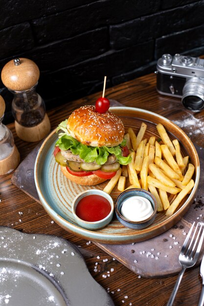 Vista lateral de hamburguesa de pollo con pepinillos y tomates servidos con papas fritas y salsas en la oscuridad