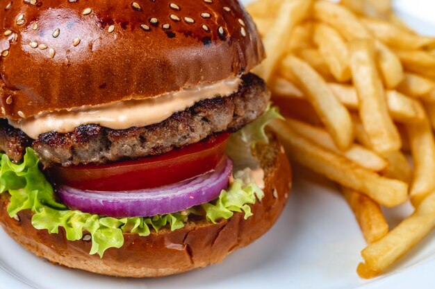 Vista lateral hamburguesa de ternera a la parrilla con salsa de tomate fresco cebolla roja lechuga entre bollos de hamburguesa y papas fritas en la mesa