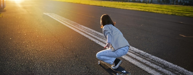 Foto gratuita vista lateral de una hermosa chica asiática en patineta montando su crucero hacia el sol en una carretera vacía