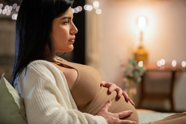 Vista lateral de una hermosa mujer embarazada sentada en la cama. Pretty Woman en cardigan tocando gran barriga. Embarazo, concepto de expectativa