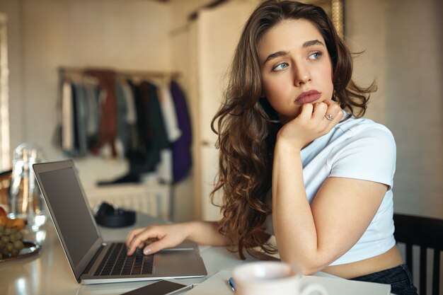 Vista lateral de la hermosa mujer joven independiente con ojos azules habiendo aburrido expresión facial triste sentado en su lugar de trabajo con portátil