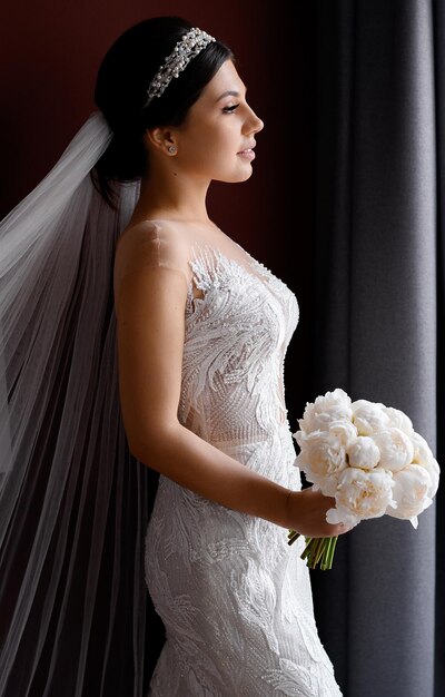 Vista lateral de una hermosa novia con peinado elegante y accesorios en la cabeza con velo largo y vestido de novia de encaje sosteniendo un ramo de peonías blancas y mirando hacia otro lado mientras posa en un estudio aislado