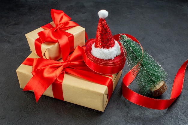 Vista lateral de hermosos regalos con cinta roja y árbol de navidad de sombrero de santa claus en mesa oscura