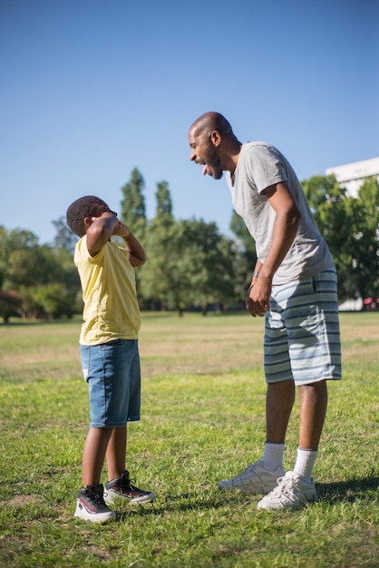 Vista lateral del hombre afroamericano y el niño haciendo muecas en el campo. Papá feliz y su lindo hijo de pie sobre un terreno cubierto de hierba mostrando lenguas el uno al otro. Concepto de ocio y momentos felices juntos