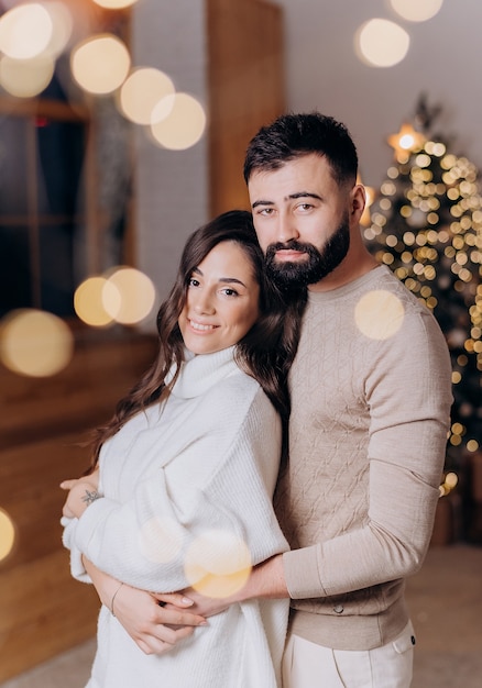 Vista lateral del hombre barbudo enamorado abraza a su novia con un suéter blanco en el fondo del árbol de Navidad