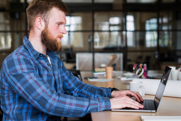 Vista lateral del hombre barbudo que lleva la camisa a cuadros azul usando la computadora portátil