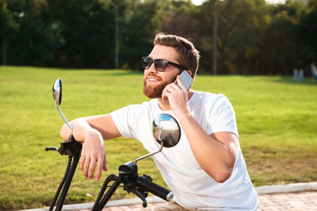 Vista lateral del hombre barbudo sonriente con gafas de sol sentado en una moto moderna al aire libre y hablando por el teléfono inteligente