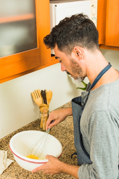 Vista lateral de un hombre batiendo los huevos con un batidor en el mostrador de la cocina