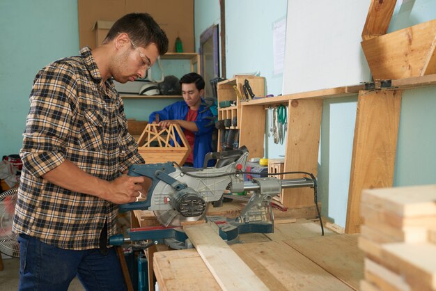 Vista lateral del hombre en camisa a cuadros aserrar madera en el taller de madera