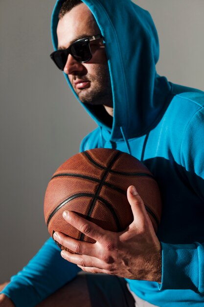 Vista lateral del hombre con capucha y gafas de sol posando mientras sostiene baloncesto