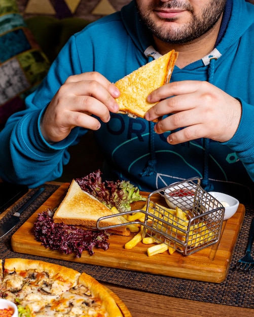 Vista lateral de un hombre comiendo clubsandwich servido con salsa de tomate y papas fritas