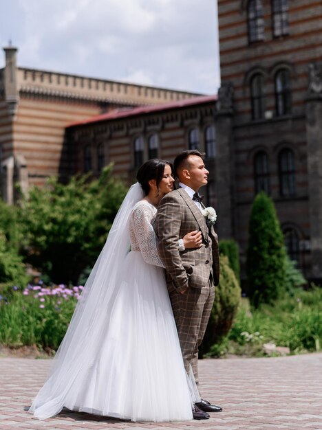 Vista lateral del hombre confiado en traje de moda manteniendo las manos en el bolsillo y mirando hacia otro lado mientras la novia en vestido de novia de pie