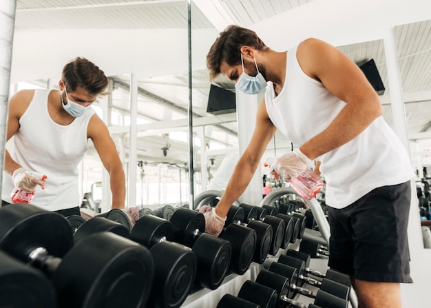 Vista lateral del hombre desinfectando equipos de gimnasio mientras usa máscara médica