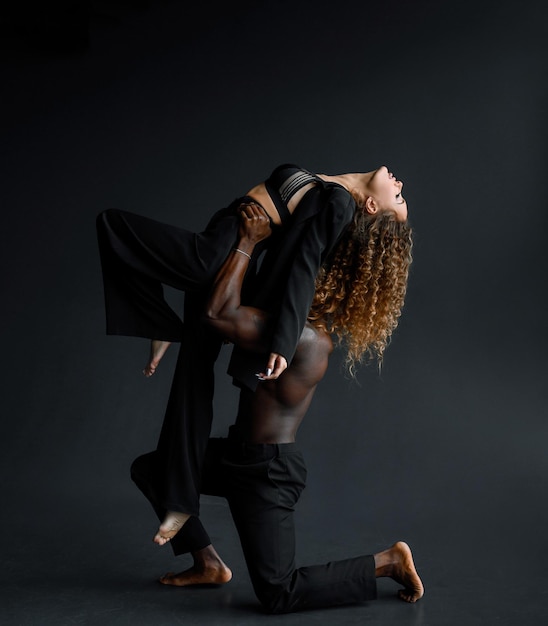 Vista lateral de un hombre fuerte de piel oscura con pantalones negros apoyado en la rodilla mientras levanta a una mujer sensual con traje negro y lencería atada mientras ensaya el baile contra el fondo negro