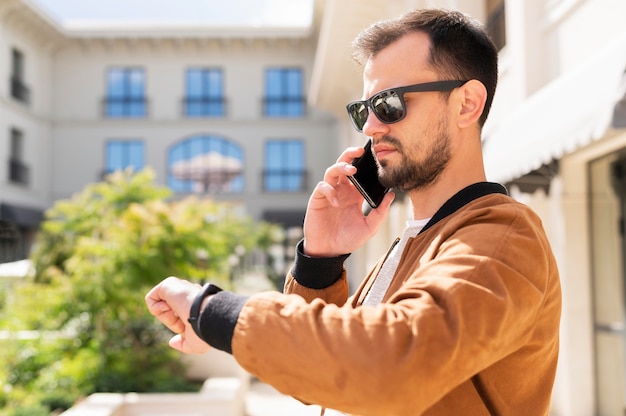 Foto gratuita vista lateral del hombre con gafas de sol que controla el tiempo mientras habla por teléfono inteligente