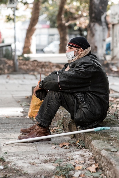 Vista lateral del hombre sin hogar al aire libre con bastón y máscara médica