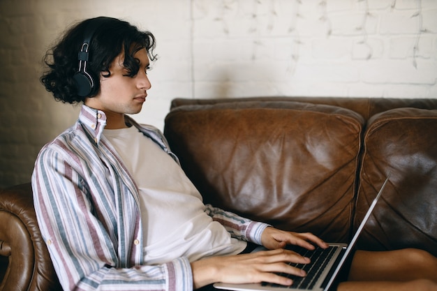 Vista lateral de un hombre joven enfocado en un sofá de cuero con una computadora portátil en su regazo usando audífonos inalámbricos escuchando música o jugando en línea, comunicándose con otros jugadores a través del chat de voz