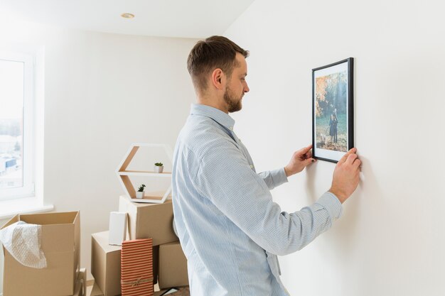 Vista lateral de un hombre joven que fija el marco de imagen en la pared en el nuevo hogar