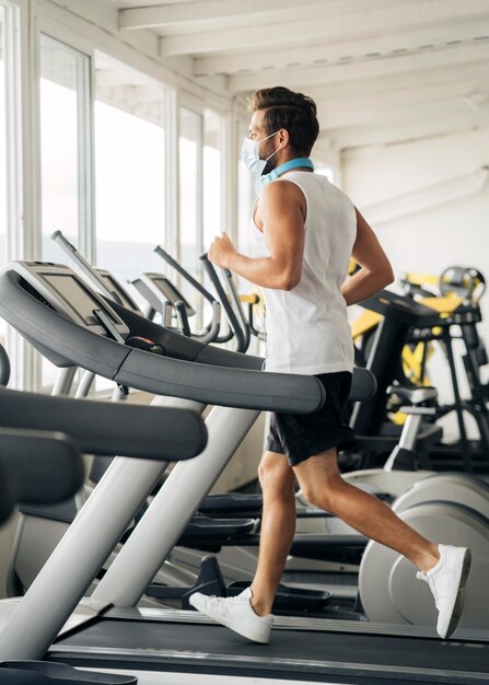 Vista lateral del hombre con máscara médica en la cinta en el gimnasio