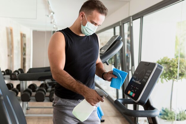 Vista lateral del hombre con máscara médica desinfectando equipos de gimnasia