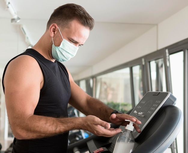 Vista lateral del hombre con máscara médica en el gimnasio con desinfectante de manos