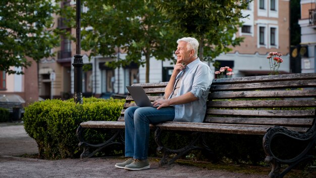 Vista lateral del hombre mayor al aire libre en un banco con un portátil