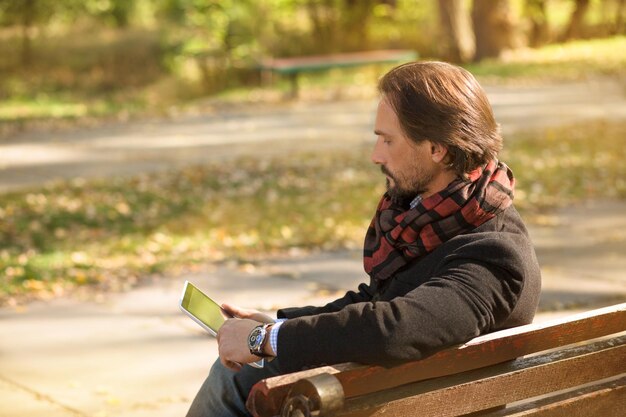 Vista lateral de un hombre de mediana edad con abrigo negro trabajando en su tablet PC. Hombre sentado en el banco al aire libre en el parque de otoño.