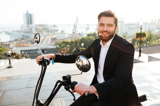 Vista lateral del hombre de negocios barbudo feliz sentado en moto