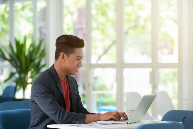 Vista lateral del hombre ocupado escribiendo en el teclado del portátil con una sonrisa