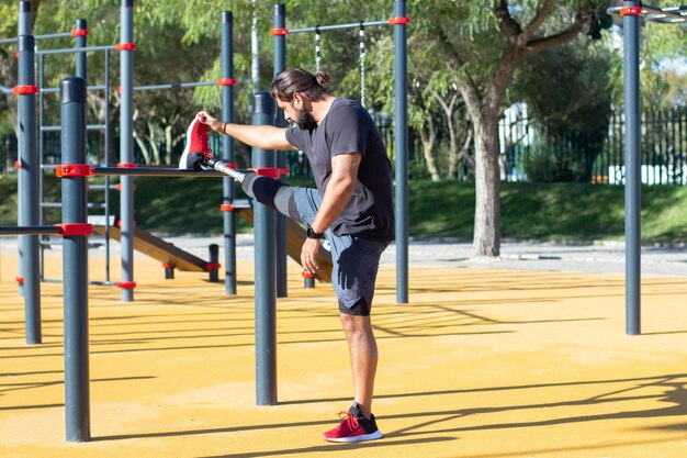 Vista lateral del hombre con prótesis de pierna entrenando al aire libre. Joven parado en el suelo sosteniendo su pierna en la barra horizontal haciendo ejercicios. Cuidado de la salud, actividades deportivas de personas con discapacidad concepto