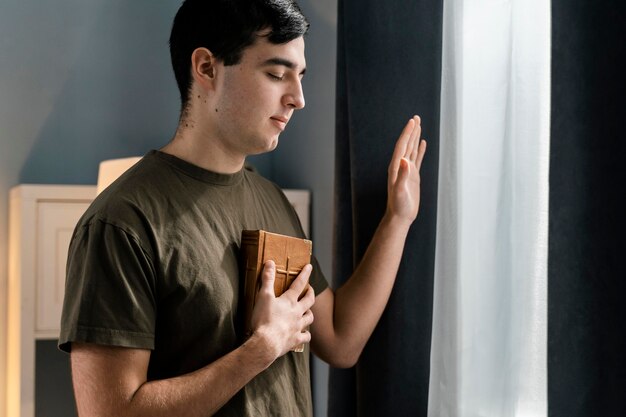 Vista lateral del hombre que sostiene la Biblia mientras está sentado junto a la ventana