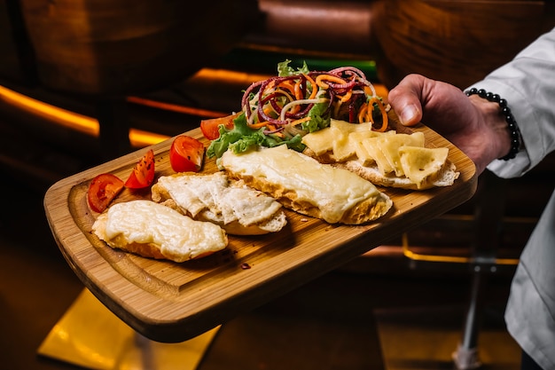 Vista lateral, un hombre sostiene una bandeja con tostadas de queso con rodajas de tomate y ensalada de verduras