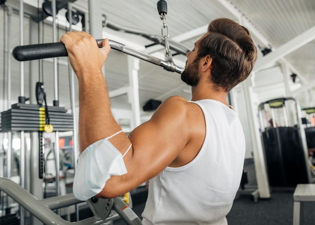 Vista lateral del hombre trabajando en el gimnasio con máscara médica en su antebrazo