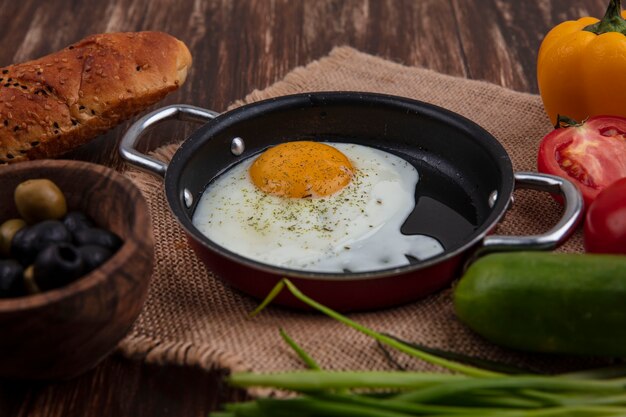 Vista lateral de huevos fritos en una sartén con cebollas verdes, aceitunas, tomates, pepinos y una hogaza de pan sobre un fondo de madera