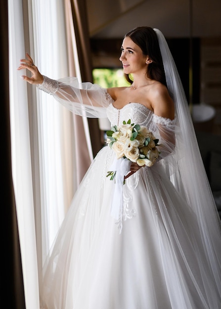 Vista lateral de una impresionante novia morena vestida con un increíble vestido blanco con hombros desnudos y un largo velo sosteniendo un ramo de flores rosas abriendo la ventana y esperando al novio durante la boda