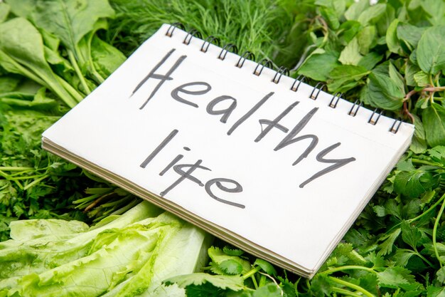 Vista lateral de la inscripción de vida sana en el cuaderno de espiral en paquetes de verduras frescas en la mesa blanca