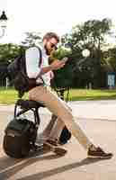 Foto gratuita vista lateral integral del hombre barbudo con gafas de sol sentado en una moto moderna al aire libre y con smartphone