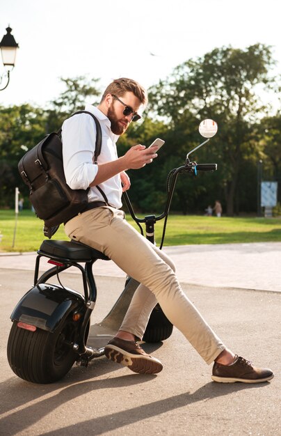 Vista lateral integral del hombre barbudo con gafas de sol sentado en una moto moderna al aire libre y con smartphone