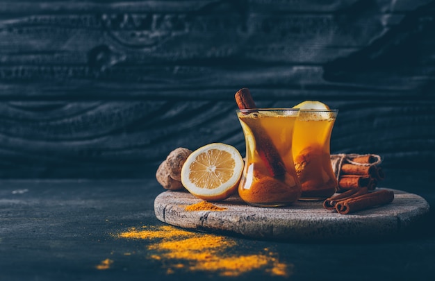 Vista lateral de jengibre en polvo en tazas de té con limón, jengibre y canela seca sobre un fondo oscuro con textura para texto