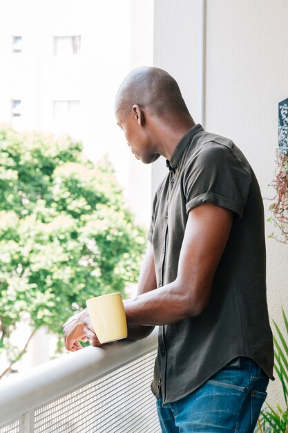 Vista lateral de un joven africano de pie en el balcón con una taza de café en la mano