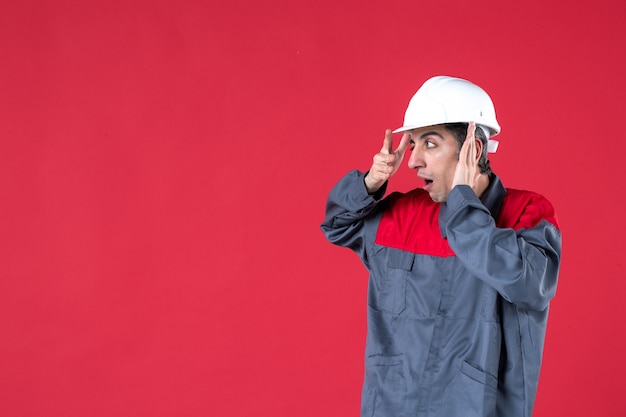 Vista lateral del joven arquitecto conmocionado emocional en uniforme con casco en pared roja aislada