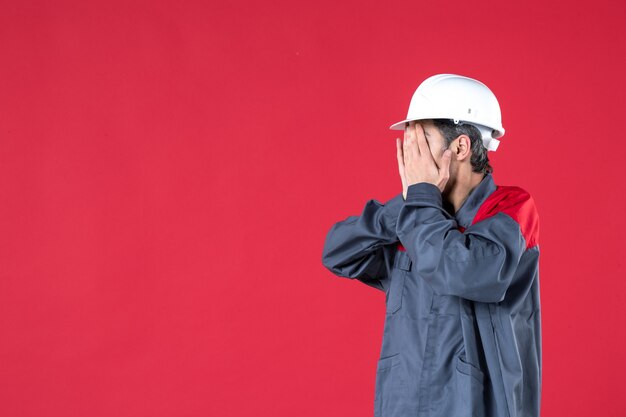 Vista lateral del joven arquitecto en uniforme con casco y cerrando su rostro con las manos en la pared roja aislada