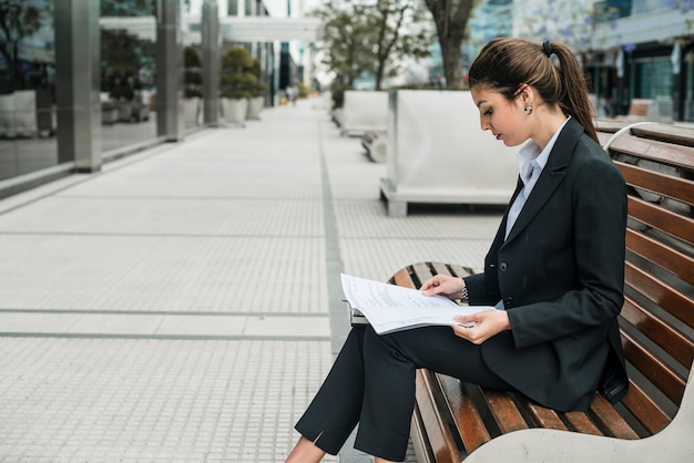 Vista lateral de una joven empresaria sentada en un banco leyendo los documentos en papel