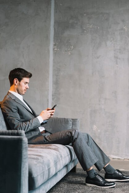 Vista lateral del joven empresario sentado en el sofá utilizando el teléfono móvil