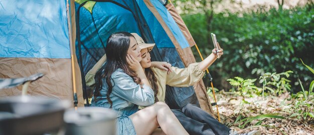 Vista lateral Joven mujer bonita asiática y su novia sentadas frente a la tienda usan el teléfono móvil para tomar una foto durante el campamento en el bosque con felicidad juntos