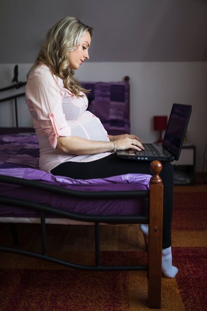 Vista lateral de una joven mujer embarazada sentada en la cama usando la computadora portátil