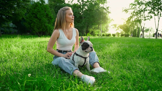 Vista lateral de la joven mujer feliz sentada sobre la hierba fresca con lindo bulldog francés blanco y marrón. Hermosa niña sonriente disfrutando del atardecer de verano, acariciar a un perro en el parque de la ciudad. Amistad humana y animal.