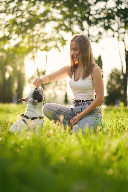 Vista lateral de la joven mujer sonriente formación bulldog francés en el parque de la ciudad. Mascota de raza pura que huele golosinas de la mano del dueño de la perra, hermosa puesta de sol de verano en el fondo. Concepto de adiestramiento animal.