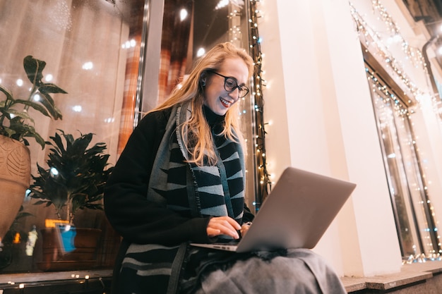 Vista lateral de la joven mujer usando una computadora portátil en la calle con la ciudad de noche