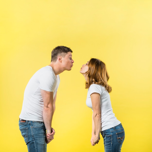 Vista lateral de una joven pareja de pie cara a cara soplando besos sobre fondo amarillo